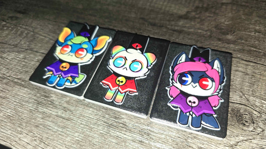 Three rectangular black rubber charms featuring colorful cartoon cat characters, displayed on a gray wood-textured surface.