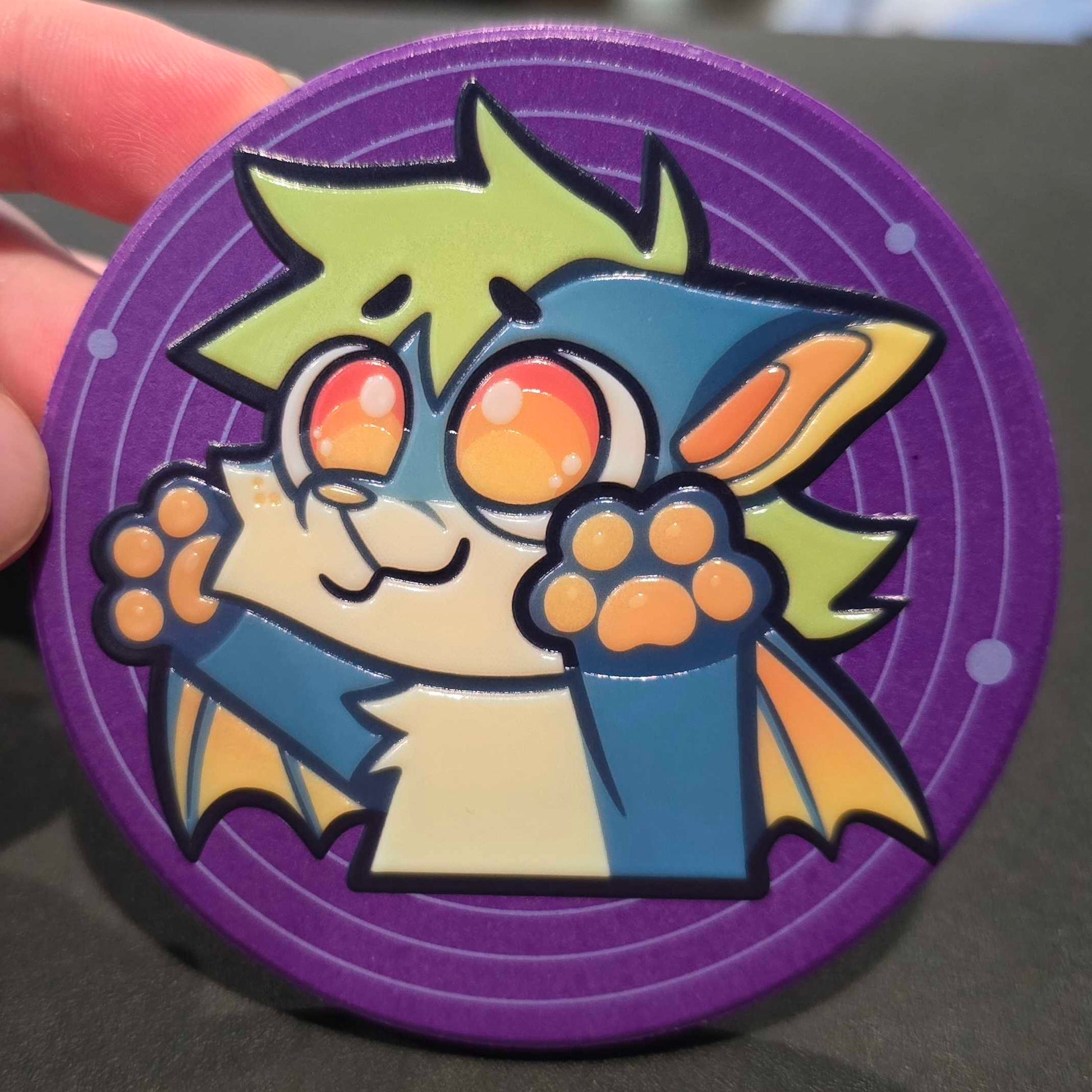 A close-up of a circular purple collectible token or medallion featuring a cartoon bat character with green hair, blue cape, and orange eyes, held by a hand against a dark background.