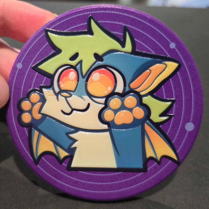 A close-up of a circular purple collectible token or medallion featuring a cartoon bat character with green hair, blue cape, and orange eyes, held by a hand against a dark background.