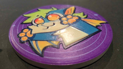 A circular purple game token or coin featuring a cartoon bat character with orange eyes, yellow wings, and a blue body, photographed on a dark surface.
