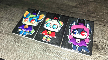 Three rectangular black rubber charms featuring colorful cartoon cat characters, displayed on a gray wood-textured surface.